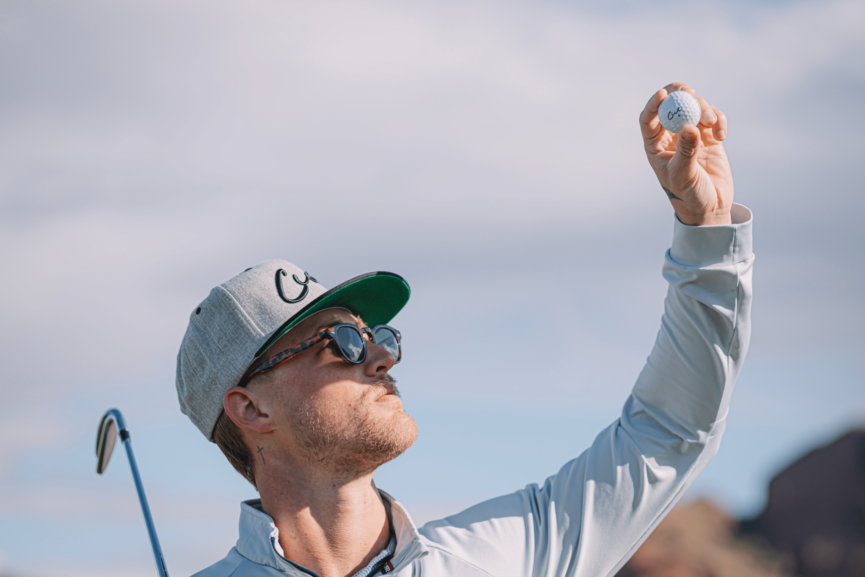 Golfer With a Cut Hat Holding a Cut Grey Golf Ball Up Towards the Sun